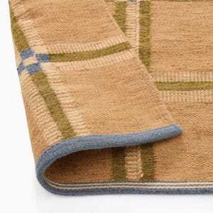 Op de afbeelding: Een geweven vloerkleed met een beige basis en een ruitpatroon van olijfgroene en lichtblauwe strepen. Het kleed heeft een dikke, getextureerde pool en een omgevouwen hoek die een lichtblauwe afgewerkte rand langs de omtrek laat zien.