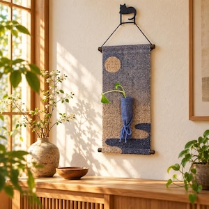Op de afbeelding: Een rechthoekige wandhanger met een getextureerd, getuft landschapsontwerp in blauw- en bruintinten met een cirkelvormige zon. Een klein blauw stoffen zakje is in het midden bevestigd, bevat een groene plant en hangt aan een zwarte katvormige haak.