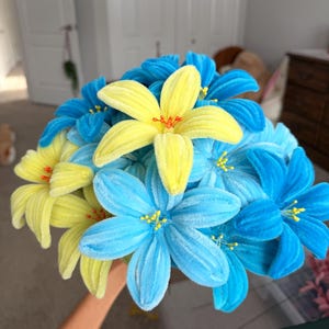 May include: A bouquet of handcrafted flowers made from fuzzy pipe cleaners in vibrant shades of blue and yellow. Each flower features six petals with a textured, velvet-like appearance and small, delicate stamens in the center of each bloom.