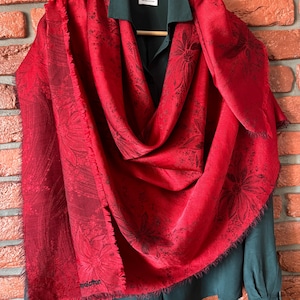 May include: A vibrant red scarf featuring a dark floral pattern and frayed edges, draped over a dark green button-down shirt. The scarf displays a repeating botanical design in black, with a visible brand label near the bottom corner.