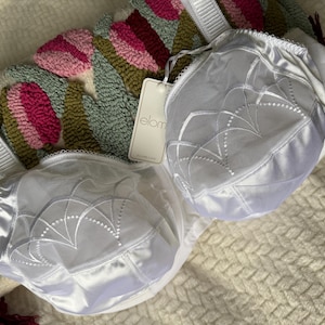 Op de afbeelding: Een witte Elomi bh met doorschijnende mesh bovencups voorzien van geborduurde waaiervormige details, effen witte onderpanelen en brede verstelbare schouderbandjes. Een rechthoekig productlabel met de merknaam is aan het midden bevestigd.
