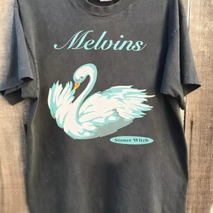 Può includere: Una t-shirt nera a maniche corte con la grafica di un cigno bianco con accenti azzurri. Il testo Melvins è stampato in un carattere corsivo azzurro sopra il cigno, e il testo Stoner Witch è stampato in un ovale azzurro sottostante.