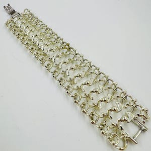 May include: Silver-toned bracelet with a unique chain design. The bracelet features multiple rows of interconnected links, each with a series of curved and rounded elements. A clasp secures the bracelet. The jewelry is displayed on a white surface.