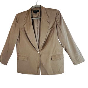 May include: A tan blazer with a single-button closure, notched lapels, and two front pockets. The blazer is hanging on a gray hanger. The fabric appears to be a smooth, woven material.