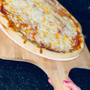 May include: A freshly baked pizza with a golden crust, topped with melted white and yellow cheese, sits on a wooden pizza peel. The pizza is ready to be served. The peel is held by a person with pink nail polish.