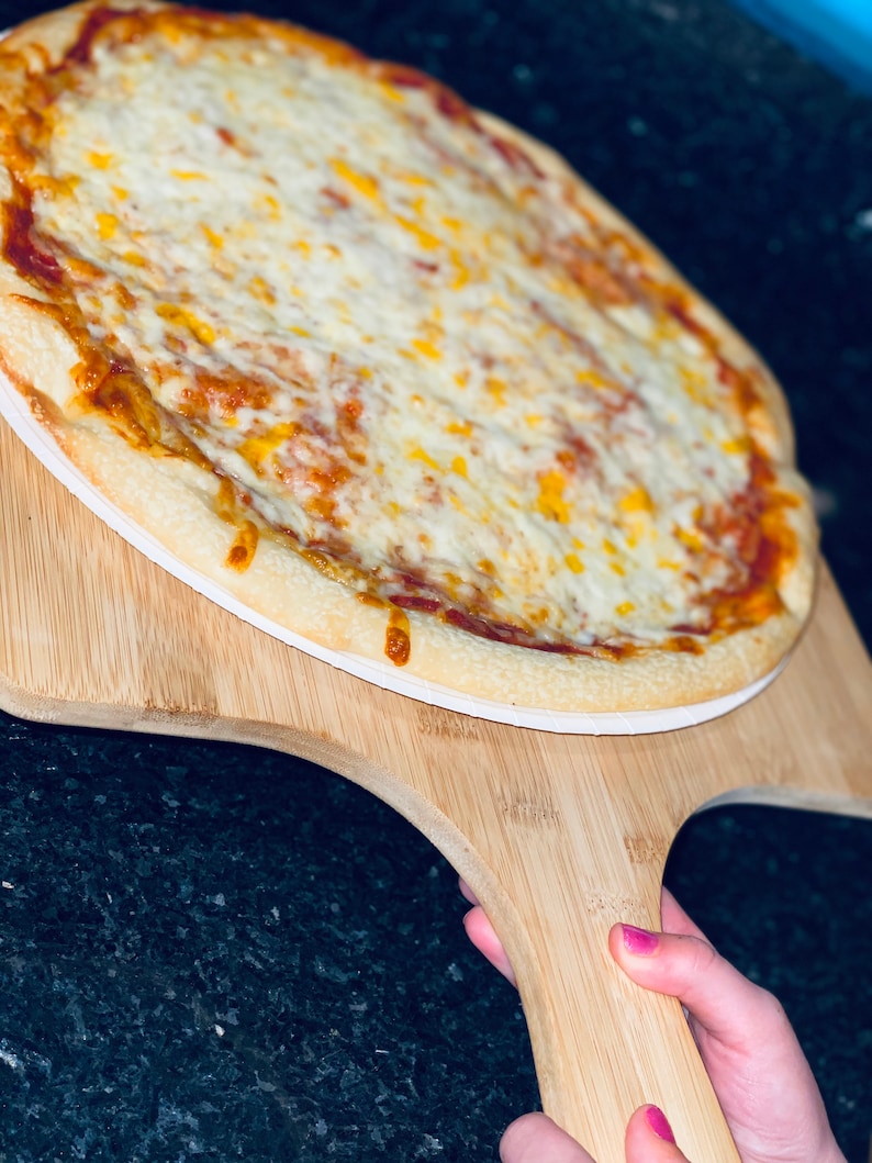 May include: A freshly baked pizza with a golden crust, topped with melted white and yellow cheese, sits on a wooden pizza peel. The pizza is ready to be served. The peel is held by a person with pink nail polish.