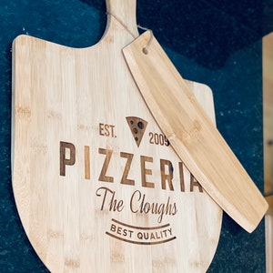 May include: A light-colored bamboo pizza peel and server set. The peel is round with a handle and the server is rectangular. The peel is engraved with "Pizzeria The Cloughs BEST QUALITY" and a cheese wedge graphic.