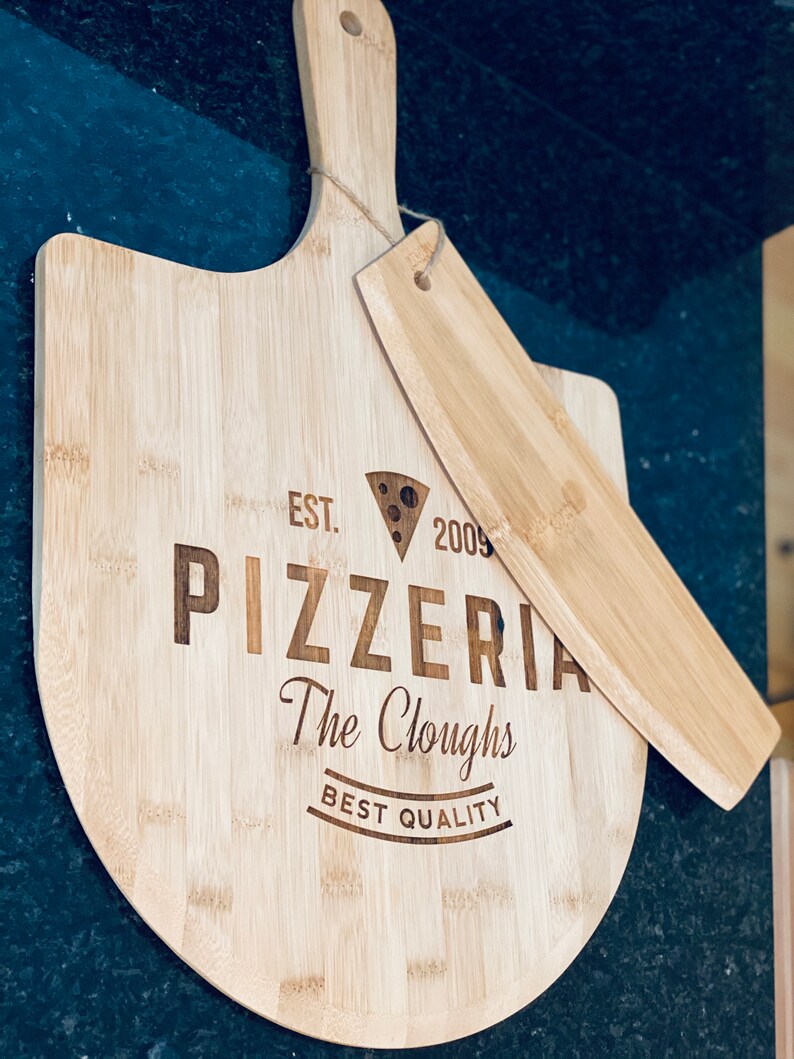 May include: A light-colored bamboo pizza peel and server set. The peel is round with a handle and the server is rectangular. The peel is engraved with "Pizzeria The Cloughs BEST QUALITY" and a cheese wedge graphic.