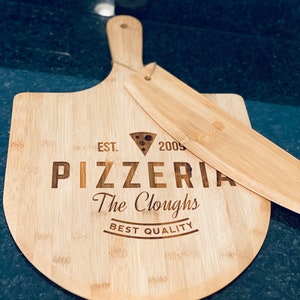 May include: A bamboo pizza peel and server set. The peel is engraved with "EST. 2005 PIZZERIA The Cloughs BEST QUALITY" and a pizza slice graphic. The server rests on the peel, both made of light-colored bamboo.
