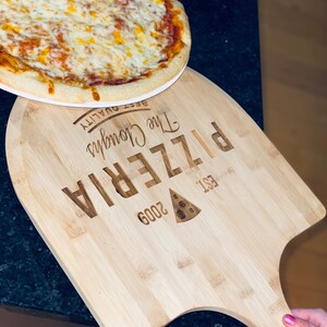 May include: A freshly baked pizza with melted cheese sits on a wooden pizza peel. The peel is engraved with "The Clough's Pizzeria" and "EST. 2009". The pizza has a golden crust and is ready to serve.