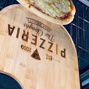 May include: A freshly baked pizza with melted cheese sits atop a wooden pizza peel. The peel is engraved with "The Cloughs Pizzeria" and "Est. 2009". The pizza is in an oven, ready to be served.