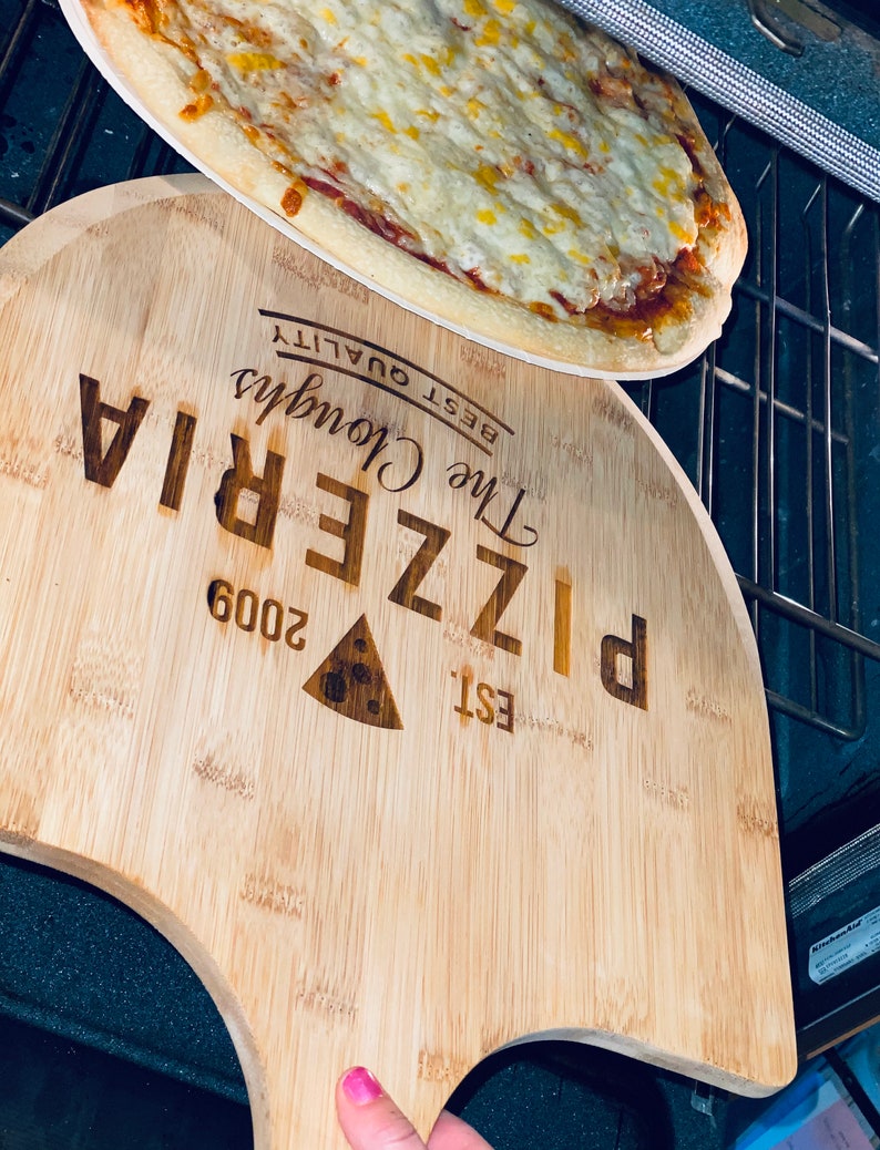 May include: A freshly baked pizza with melted cheese sits atop a wooden pizza peel. The peel is engraved with "The Cloughs Pizzeria" and "Est. 2009". The pizza is in an oven, ready to be served.