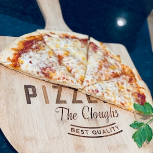 May include: A round wooden pizza peel with a handle, holding a sliced cheese pizza. The peel is engraved with the word "PIZZA", the name "The Cloughs", and "BEST QUALITY". A sprig of green herbs is on the side.