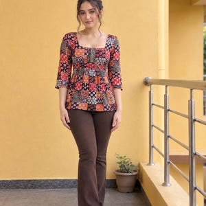 Op de afbeelding: Een top met driekwartmouwen, een vierkante halslijn en een veelkleurig patchworkpatroon in rood-, zwart- en oranjetinten, gecombineerd met een effen donkerbruine broek met wijde pijpen en witte sneakers voor een complete outfit.