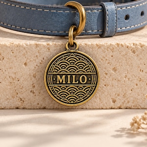 May include: A blue leather pet collar with white stitching and a circular antique brass tag. The tag features a repeating wave pattern and the name MILO engraved in the center. The metal hardware includes a sturdy ring for attachment.