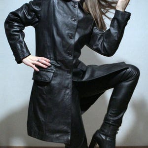 May include: A black leather trench coat featuring a classic collar, a front button closure, and two side pockets with flaps. The coat is paired with matching black leather trousers and black leather heeled boots, creating a coordinated ensemble.