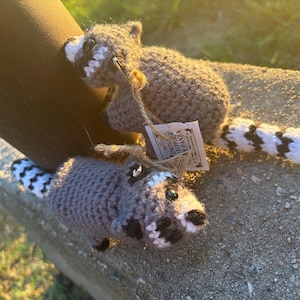 May include: Two crocheted raccoon plush toys made with grey, white, and black yarn. Each raccoon features a striped tail, black eye patches, and a small tag attached with twine that reads Emotional Support Raccoon. The toys have a soft, textured finish.