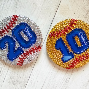 May include: Two round, bedazzled ornaments. One is silver with red accents and the number 20 in blue with the name "Beau". The other is gold with red accents and the number 10 in blue with the name "Dakota".