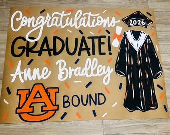 May include: A graduation sign on brown paper with the words "Congratulations GRADUATE! Anne Bradley" in white and black. An illustration of a graduation gown and cap with "2026" is on the right. The Auburn University logo is at the bottom.