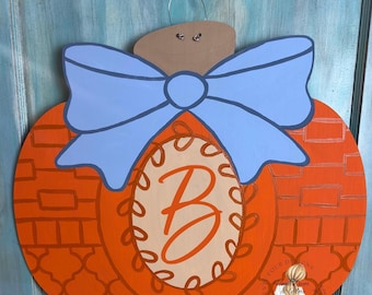 May include: Orange wooden pumpkin-shaped door hanger with a light blue bow and a beige oval in the center. The oval features the letter "B" in an orange script font. The pumpkin has a brick pattern.