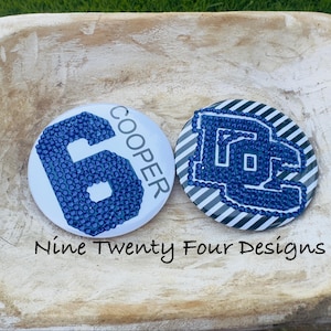 May include: Two round, white buttons with blue designs. One button features a blue number 6 with the name "COOPER" in blue. The other button has a blue logo on a striped background. The text "Nine Twenty Four Designs" is below the buttons.
