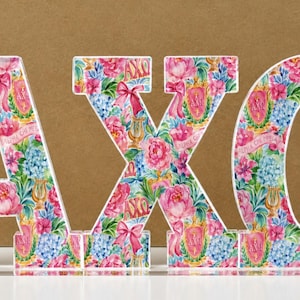 May include: Three-dimensional letters spelling AXO, featuring a vibrant floral pattern with pink peonies, blue hydrangeas, gold lyres, pink ribbons, and the Greek letters Alpha Chi Omega printed on a white background, mounted on a clear base.