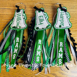 May include: Three personalized cheerleading keychains with green, black, and white ribbons. Each keychain features a megaphone design with a name and the word "RAMS". The keychains are embellished with various ribbons and beads.
