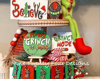 May include: A tiered tray decorated for Christmas with a Grinch theme. The tray features a green Grinch gnome, a red and white candy cane, a sign that says "Believe", a round sign that says "Merry Grinchmas", a sign that says "Grinch Mode", and a banner that says "Nine Twenty Four Designs".