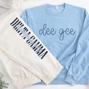 May include: A light blue sweatshirt with the cursive text "dee gee" and cream-colored sweatpants with "DELTA GAMMA" printed vertically in navy blue. The sweatpants also have the text "Established 1873".
