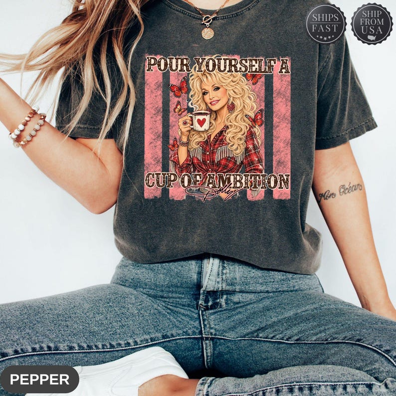 Op de afbeelding: Een antracietgrijs T-shirt met korte mouwen en een afbeelding van een blonde vrouw die een koffiemok met een hart vasthoudt. De tekst luidt "POUR YOURSELF A CUP OF AMBITION" met rode vlinders en roze verticale strepen op de achtergrond.