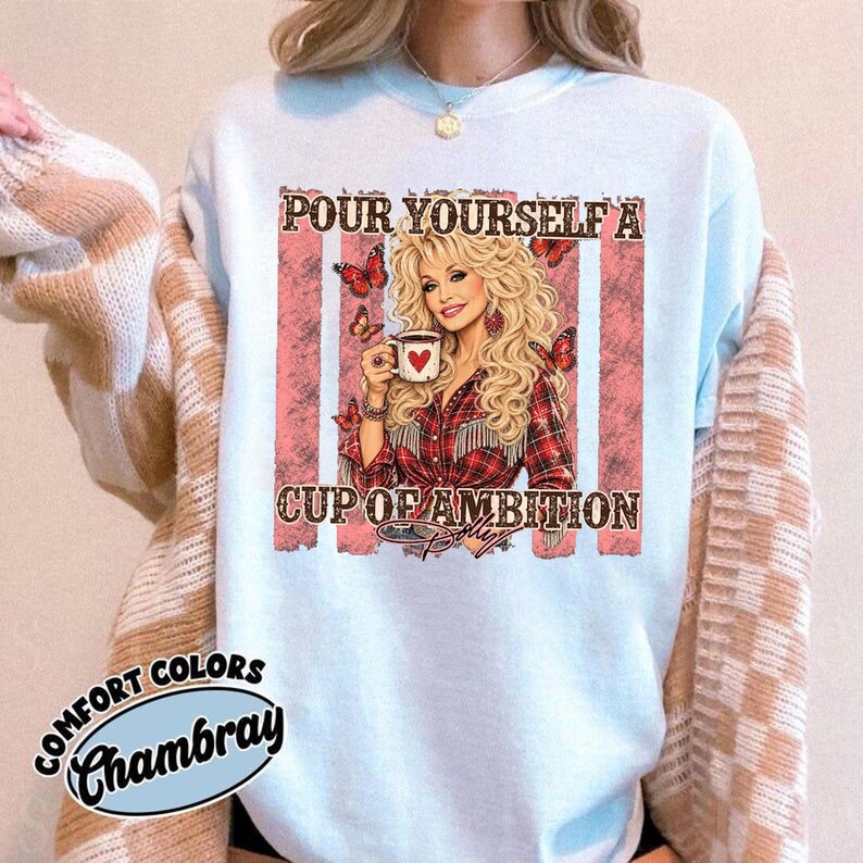 Op de afbeelding: Een lichtblauw sweatshirt met ronde hals en een grafische print van een blonde vrouw die een koffiemok met een hart vasthoudt. De tekst luidt "POUR YOURSELF A CUP OF AMBITION" met vlinders en verticale roze strepen. Een "COMFORT COLORS CHAMBRAY" label is zichtbaar.