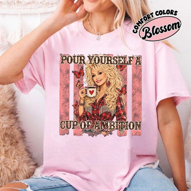 Op de afbeelding: Een lichtroze t-shirt met een afbeelding van een blonde vrouw in een rood geruit overhemd die een koffiemok met een hartje vasthoudt. De tekst luidt "POUR YOURSELF A CUP OF AMBITION" met vlinders rond de figuur. Het t-shirt heeft een ronde hals.