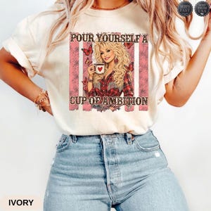 Op de afbeelding: Een ivoorkleurig T-shirt met korte mouwen en een grafische print van een blonde vrouw die een koffiemok vasthoudt. De tekst luidt "POUR YOURSELF A CUP OF AMBITION" met vlinderaccenten en roze verticale strepen in het ontwerp.