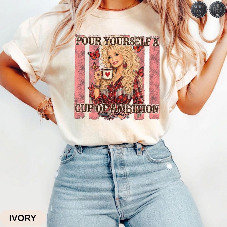 Op de afbeelding: Een ivoorkleurig T-shirt met korte mouwen en een grafische print van een blonde vrouw die een koffiemok vasthoudt. De tekst luidt "POUR YOURSELF A CUP OF AMBITION" met vlinderaccenten en roze verticale strepen in het ontwerp.