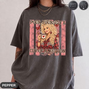 Op de afbeelding: Een antracietgrijs t-shirt met korte mouwen en een grafische print van een blonde vrouw in een rood geruit overhemd die een witte mok met een hart vasthoudt. De tekst luidt POUR YOURSELF A CUP OF AMBITION. Rode verticale strepen en vlinders.