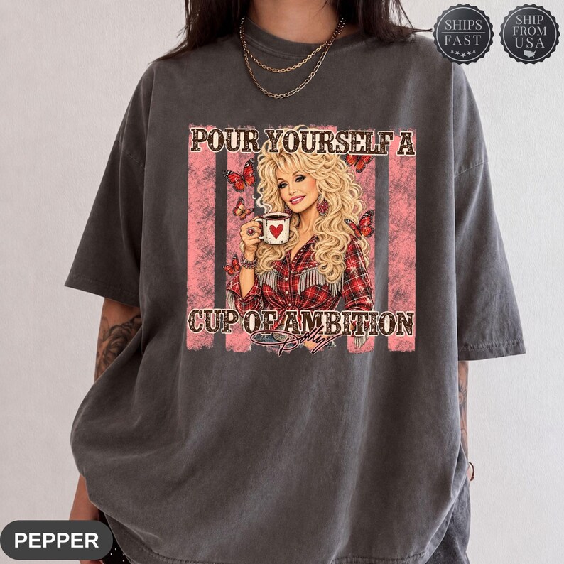 Op de afbeelding: Een antracietgrijs t-shirt met korte mouwen en een grafische print van een blonde vrouw in een rood geruit overhemd die een witte mok met een hart vasthoudt. De tekst luidt POUR YOURSELF A CUP OF AMBITION. Rode verticale strepen en vlinders.