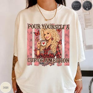 Op de afbeelding: Een ivoorkleurig t-shirt met een afbeelding van een blonde vrouw in een rood geruit overhemd die een koffiemok met een hart vasthoudt. De tekst luidt "POUR YOURSELF A CUP OF AMBITION" met vlinderaccenten op een roze, verweerde achtergrond.