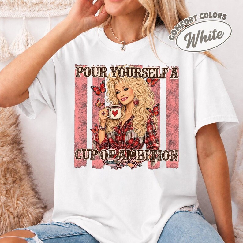 Op de afbeelding: Een wit t-shirt met een grafische print van een blonde vrouw die een koffiemok met een hartje vasthoudt. De tekst luidt "POUR YOURSELF A CUP OF AMBITION" en "DOLLY". Het ontwerp bevat rode vlinders en een roze verweerd achtergrondpatroon.