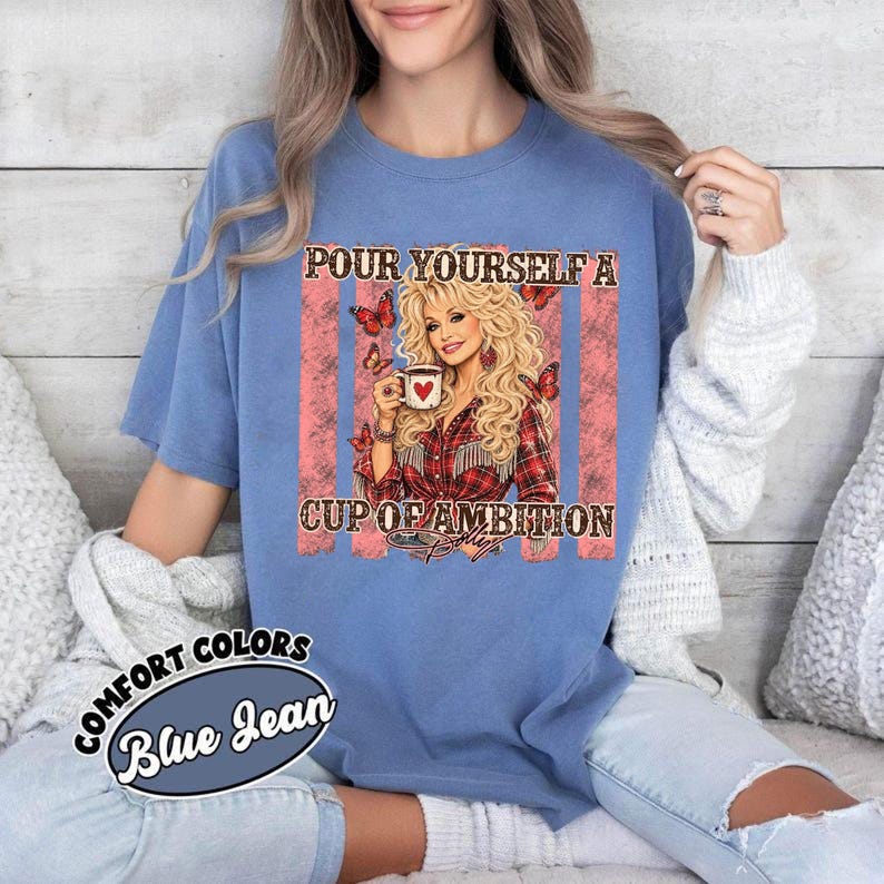 Op de afbeelding: Een blauw spijkerkleurig t-shirt met een afbeelding van een blonde vrouw die een koffiemok met een rood hart vasthoudt. De tekst luidt "POUR YOURSELF A CUP OF AMBITION" met vlinderillustraties op een achtergrond van roze en witte strepen.