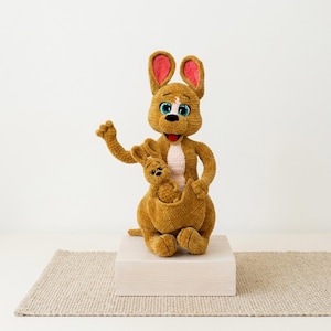 Peut inclure: Un jouet en peluche kangourou crocheté marron avec une tache ventrale rose clair, de grands yeux bleus et des oreilles doublées de rose. Le kangourou a un bras levé et un petit bébé kangourou niché dans sa poche ventrale, avec des détails cousus.