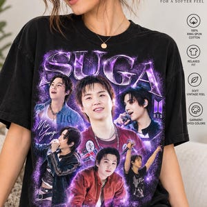 Può includere: Una t-shirt nera a maniche corte con una stampa grafica dell'artista Suga dei BTS. Il design include il testo SUGA, Min Yoongi e BTS, circondato da effetti di luce viola e diverse immagini dell'artista. La maglietta ha una vestibilità rilassata.