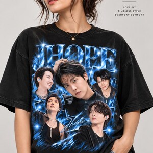 Peut inclure: Un t-shirt noir à manches courtes avec une impression graphique de l'artiste J-Hope dans diverses poses, des effets d'étoiles bleues et le texte JHOPE. Le t-shirt porte l'inscription COMFORT COLORS PREMIUM GARMENT DYED TEE SOFT FIT TIMELESS STYLE EVERYDAY COMFORT.