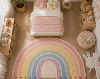 Pode incluir: Um tapete semicircular com um padrão de arco-íris em tons pastel, composto por faixas concêntricas de rosa, amarelo, verde, azul e roxo. O tapete é rematado com pompons de lã em cores pastel coordenadas ao longo da borda inferior reta.