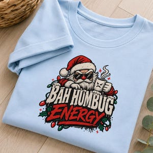 Könnte beinhalten: Ein hellblaues T-Shirt mit einer Grafik des Weihnachtsmanns, der eine Sonnenbrille trägt und eine dampfende Tasse hält. Der Text "BAH HUMBUG ENERGY" ist in weißer und roter Schrift gedruckt, umgeben von Weihnachtslichtern und Stechpalmen.