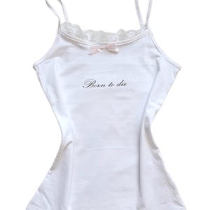 Pode incluir: Uma camisola branca com alças finas, um delicado acabamento em renda ao longo do decote e um pequeno laço rosa claro centrado no peito. O texto "Born to die" está impresso numa fonte cursiva preta na parte da frente da peça.