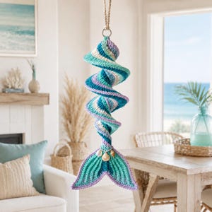 May include: A spiral-shaped crochet hanging decoration featuring a mermaid tail at the bottom. The piece is crafted with yarn in shades of teal, turquoise, white, and lavender. Three small natural seashells are attached to the top of the tail.