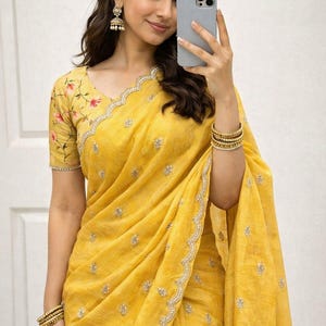 May include: A vibrant yellow saree featuring a scalloped border with intricate silver embroidery and small floral motifs. The matching blouse displays a delicate pink and green floral print on the sleeves, complemented by gold-toned bangles.
