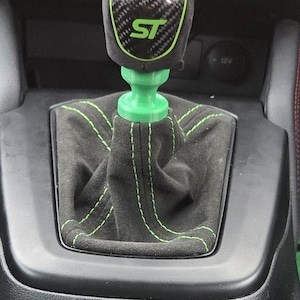 May include: A car gear shift knob featuring a carbon fibre pattern with a green ST logo and green trim. The gear shift boot is made of black fabric with green stitching, connected to a bright green base, set within a black centre console.