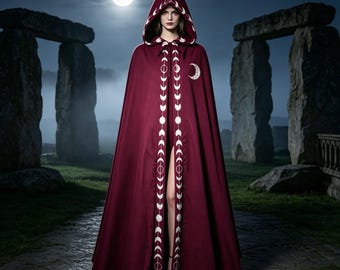 Può includere: Un mantello con cappuccio color bordeaux profondo che arriva fino a terra, caratterizzato da un motivo ricamato bianco delle fasi lunari lungo l'apertura frontale e il bordo del cappuccio, con un emblema a mezzaluna sul petto e chiusura.