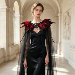 May include: A black satin floor-length dress featuring a sheer black tulle cape overlay. The neckline is adorned with a decorative black lace collar embellished with red and black feathers and a central red gemstone pendant with hanging chains.